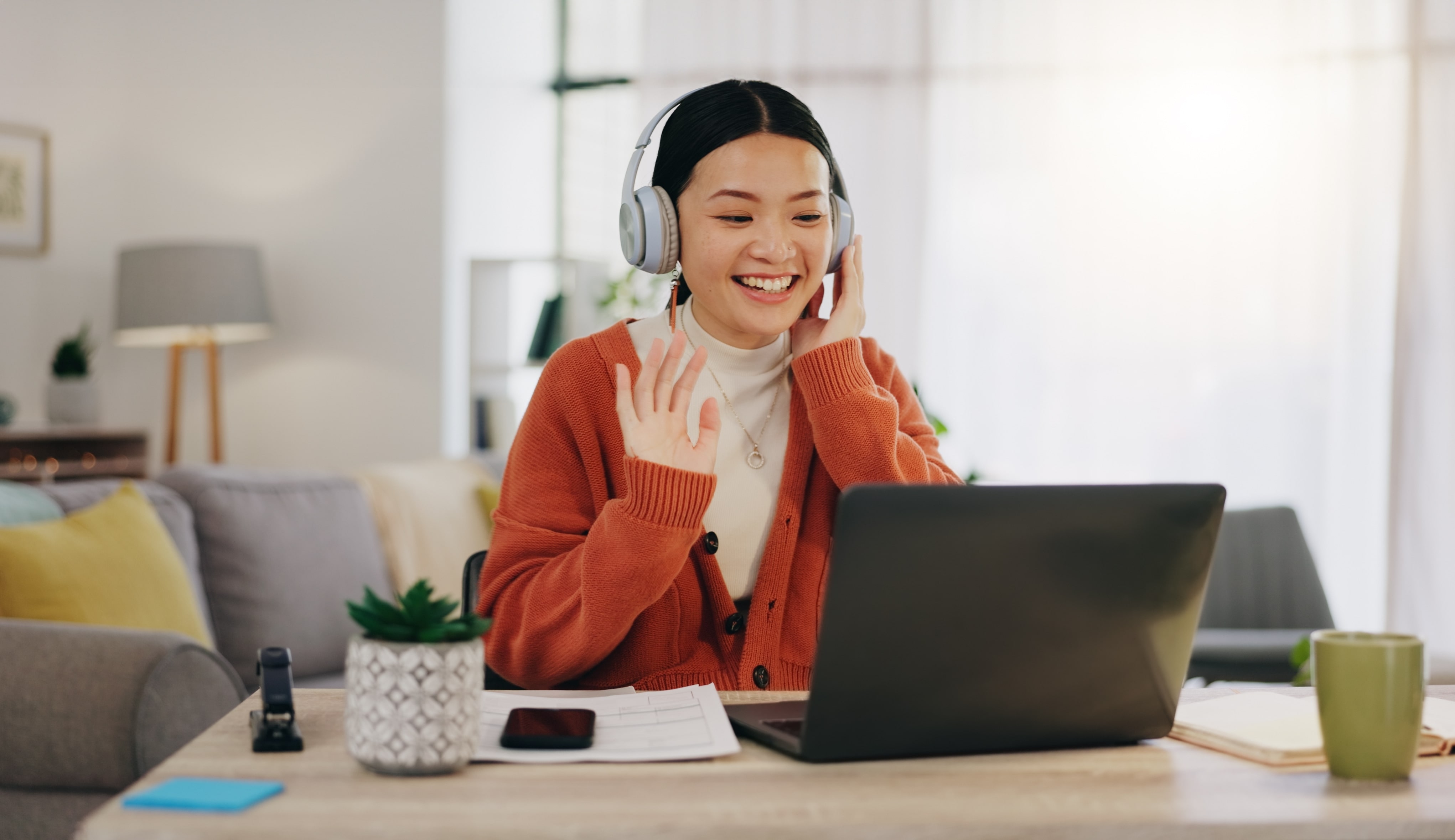 Imagem de uma jovem mulher sentada em uma mesa, interagindo com alguém através de uma videochamada em seu notebook. Ela está usando um fone de ouvido e sorrindo, enquanto acena com a mão direita. Ao fundo, é possível ver um ambiente de sala de estar. 