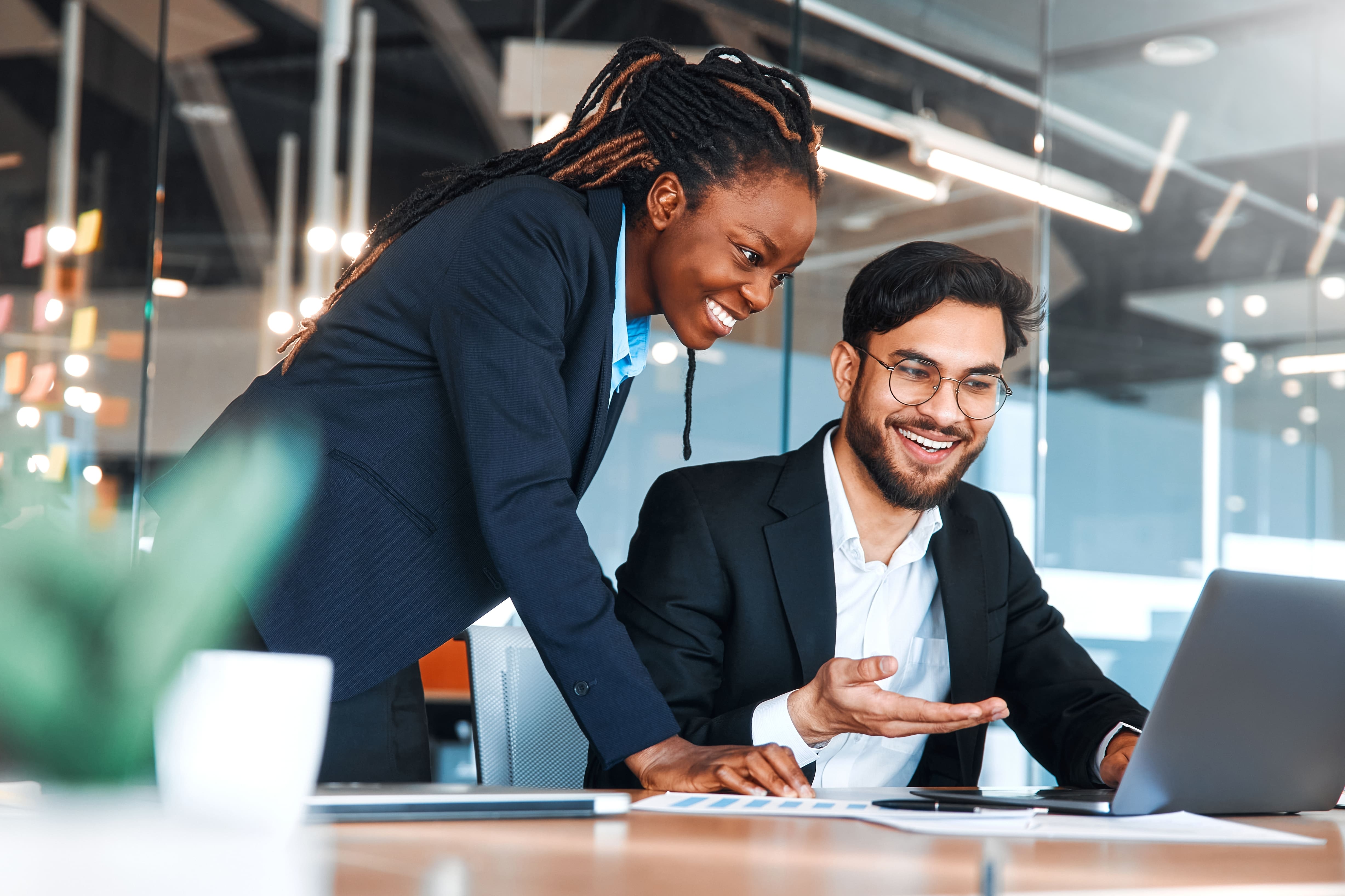 Dois profissionais, um homem e uma mulher, discutindo estratégias de trabalho em um ambiente de escritório moderno. Ambos estão sorrindo e focados na tela de um laptop, representando um ambiente colaborativo em uma empresa multinacional.